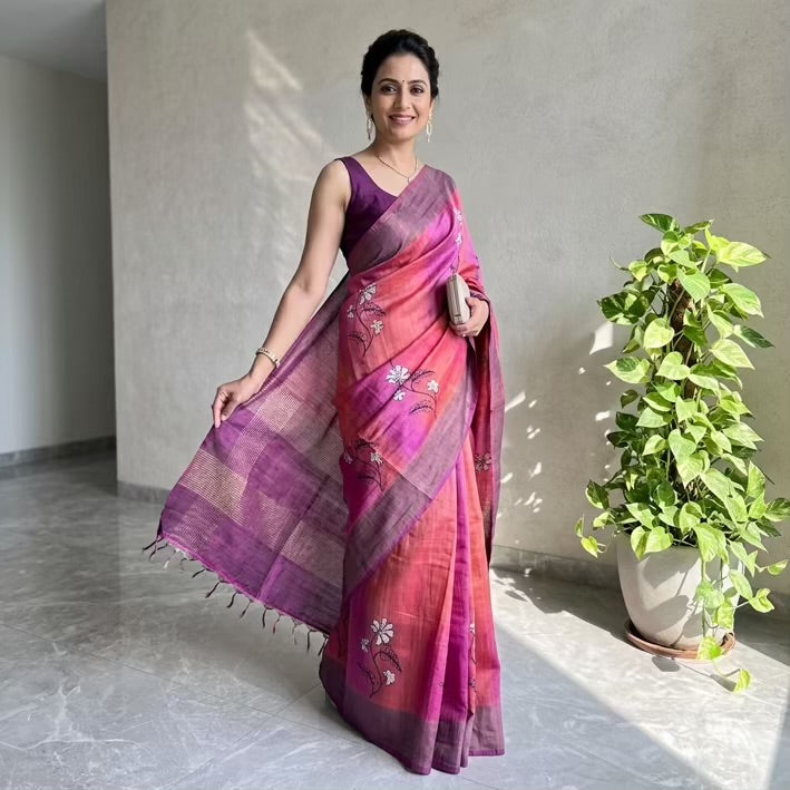 A woman models a pink and purple striped silk saree with white floral embroidery, paired with a solid purple sleeveless blouse, standing in a sunlit room next to a potted green plant.