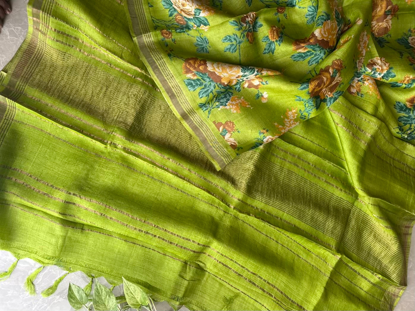A close-up of a bright lime green saree or fabric with a floral print and woven stripes. The fabric features a mix of solid lime green, a patterned section with orange and blue flowers, and a border with gold-toned woven details.