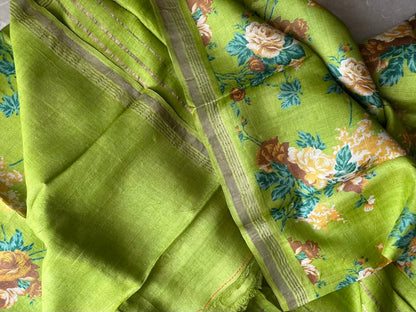 A close-up of a lime green fabric, likely a saree, featuring a floral print with yellow and white roses, blue leaves, and a beige-gold zari border.