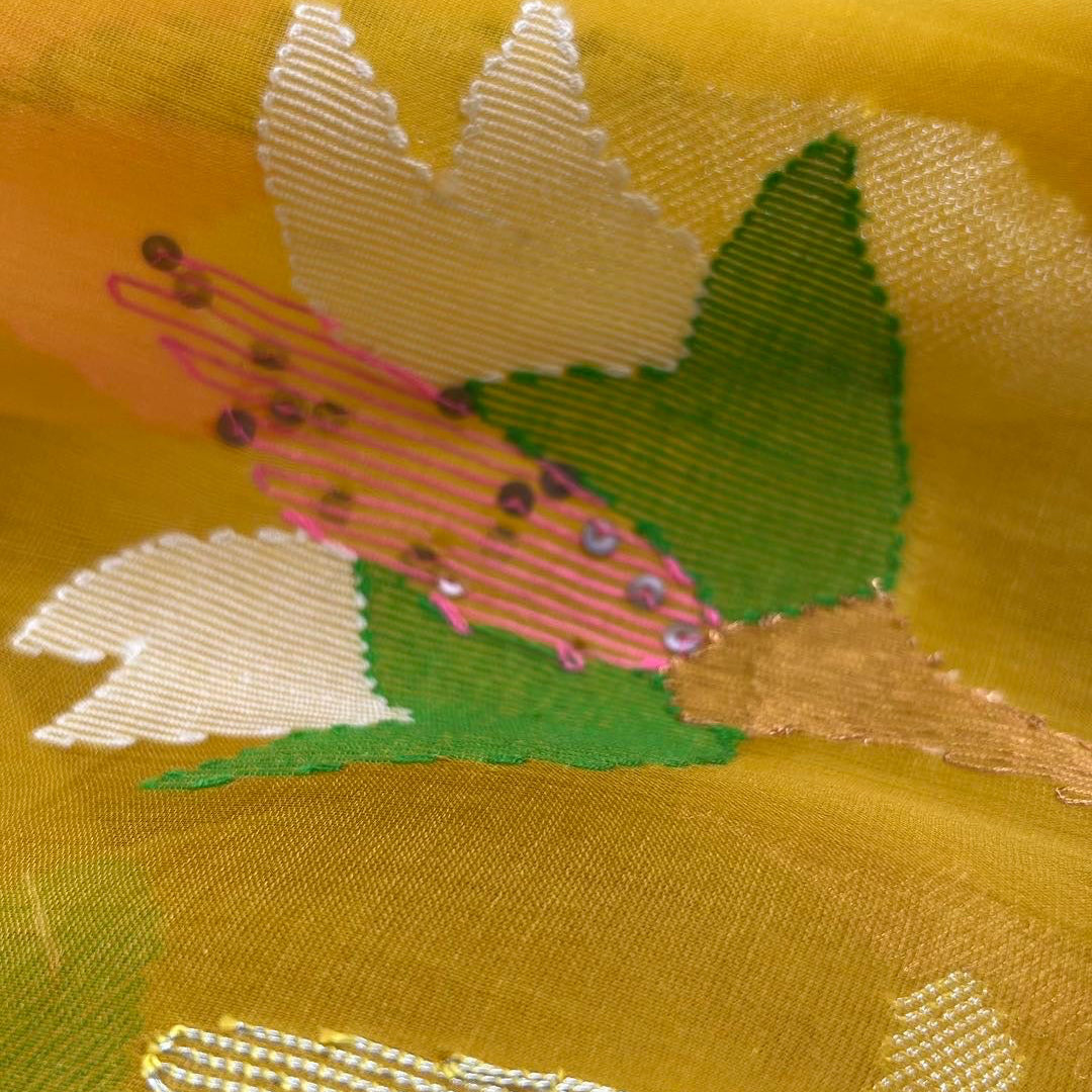 Close-up of a yellow sheer fabric with woven floral motifs in white, green, and pink, accented with small sequins.