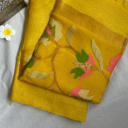 A folded yellow saree with a floral print in pink, green, and white, displayed on a white fabric surface next to a small flower and a decorative box.