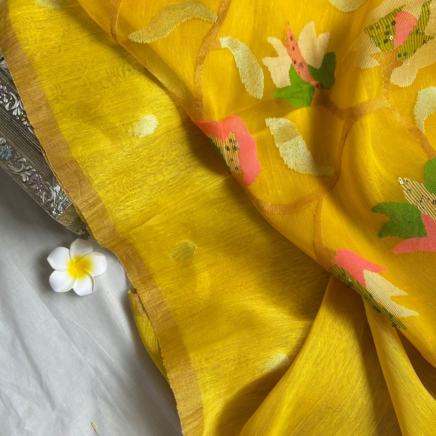 A close-up of a bright yellow fabric, likely a saree or scarf, featuring hand-woven floral motifs in pink, green, and cream, with a gold border. A small white flower is placed on a white surface next to the fabric.