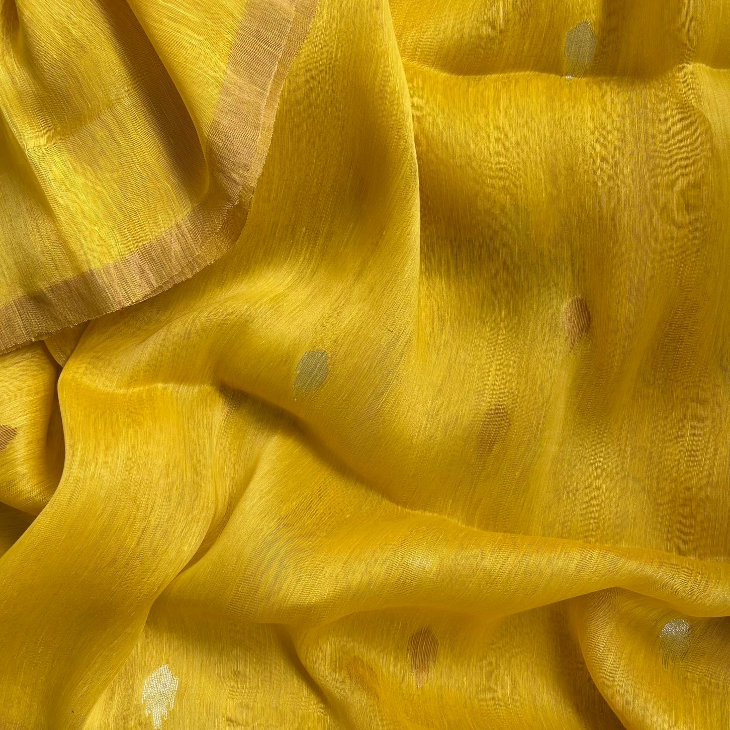 A close-up of draped yellow fabric with subtle metallic gold and silver woven accents, showing its soft texture and folds.