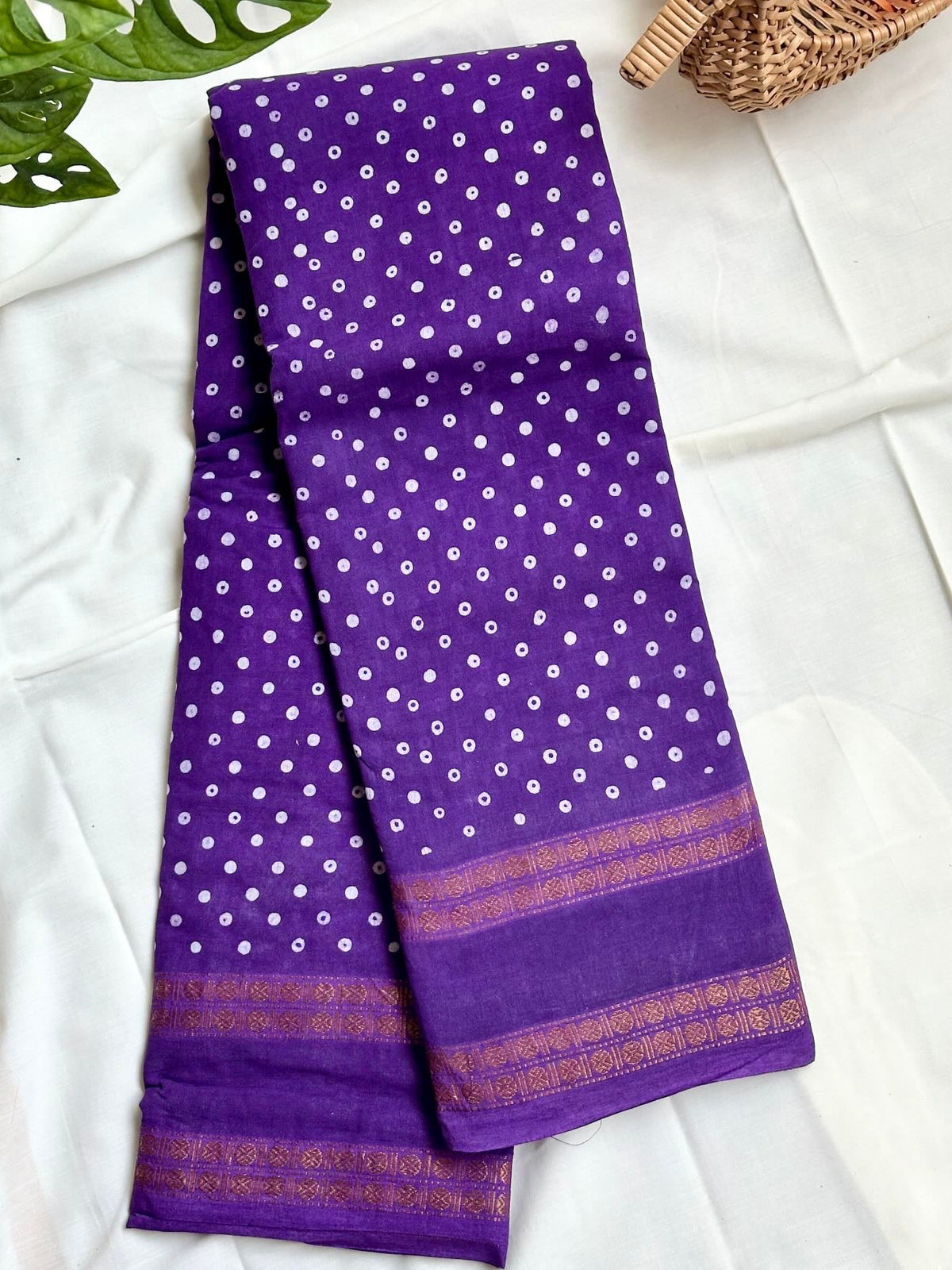 A folded purple saree with white tie-dye dots and a gold patterned border, displayed on a white fabric background with decorative green leaves and a woven basket.