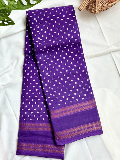A folded purple saree with white tie-dye dots and a gold patterned border, displayed on a white fabric background with decorative green leaves and a woven basket.