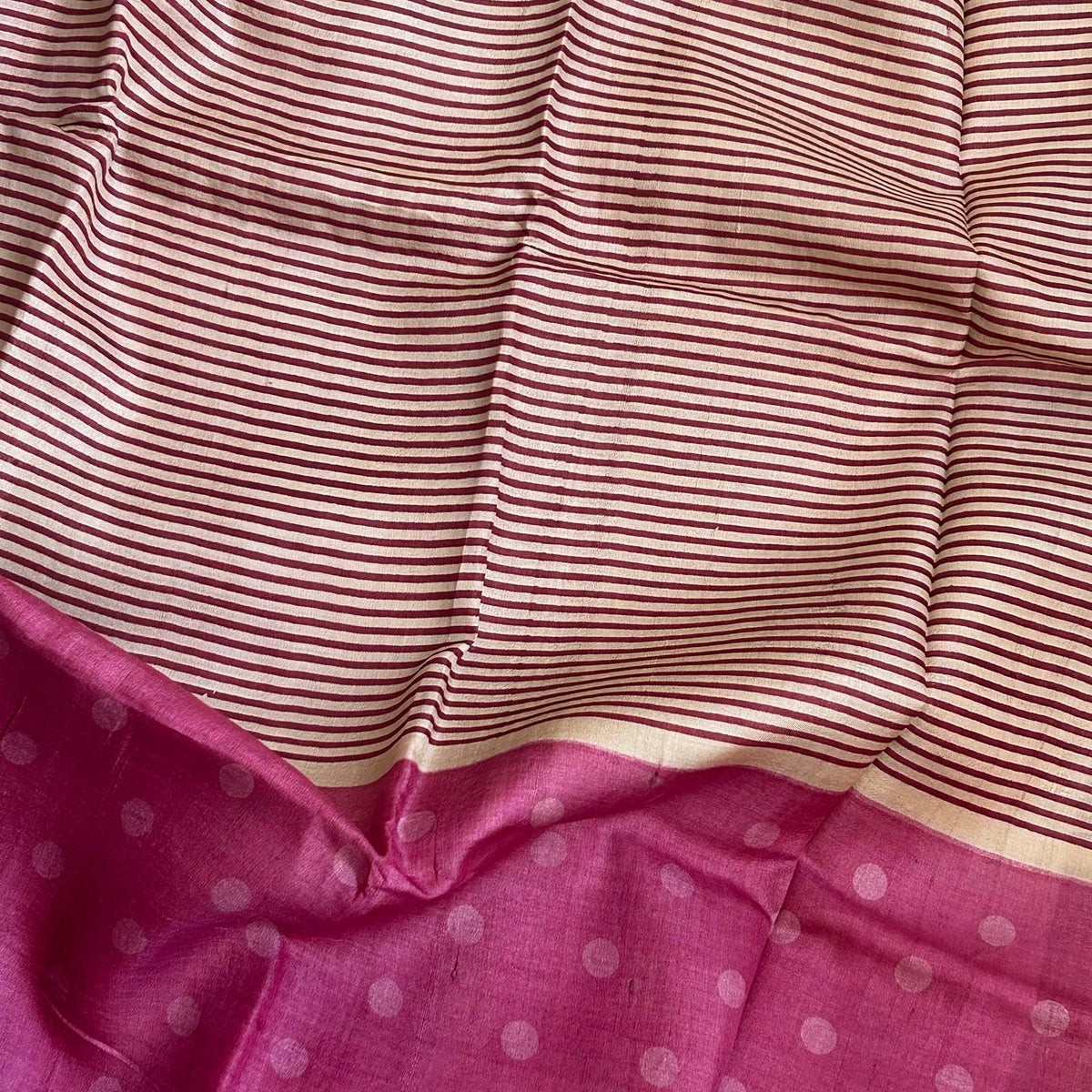 A close-up of a striped Tussar saree, featuring thin dark red horizontal stripes on a light beige base, with a solid magenta-pink border at the bottom decorated with small white polka dots.